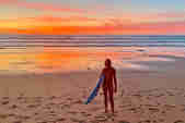 A female surfer holding a surfboard under her arm, standing on the beach with back to the shore. Gentle small waves and the colours of an orange and red sunset spread across the sky