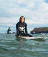 A person sits on a surfboard smiling at the camera in the sea at Watergate Bay