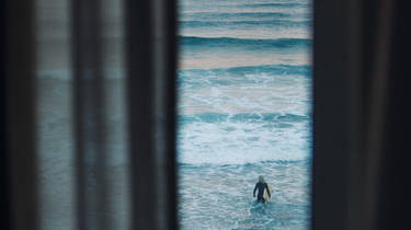 Curtains framing the photo with a view of a surfer walking into the sea with their surf board