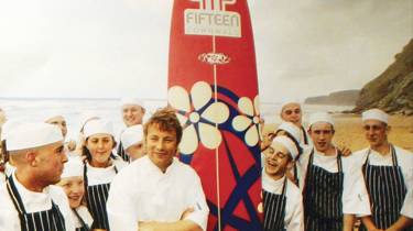 Jamie Oliver on the beach at Watergate Bay with a surfboard and apprentices