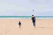 Father and son running on the sand towards the sea with a kite surfer in the background